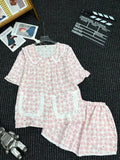 A two-piece pajama set laid flat on a dark textured surface, featuring a pink and white gingham pattern with small black motifs, white ruffled trim, and a ruffled collar on the short-sleeve top. The matching shorts are placed beside it.