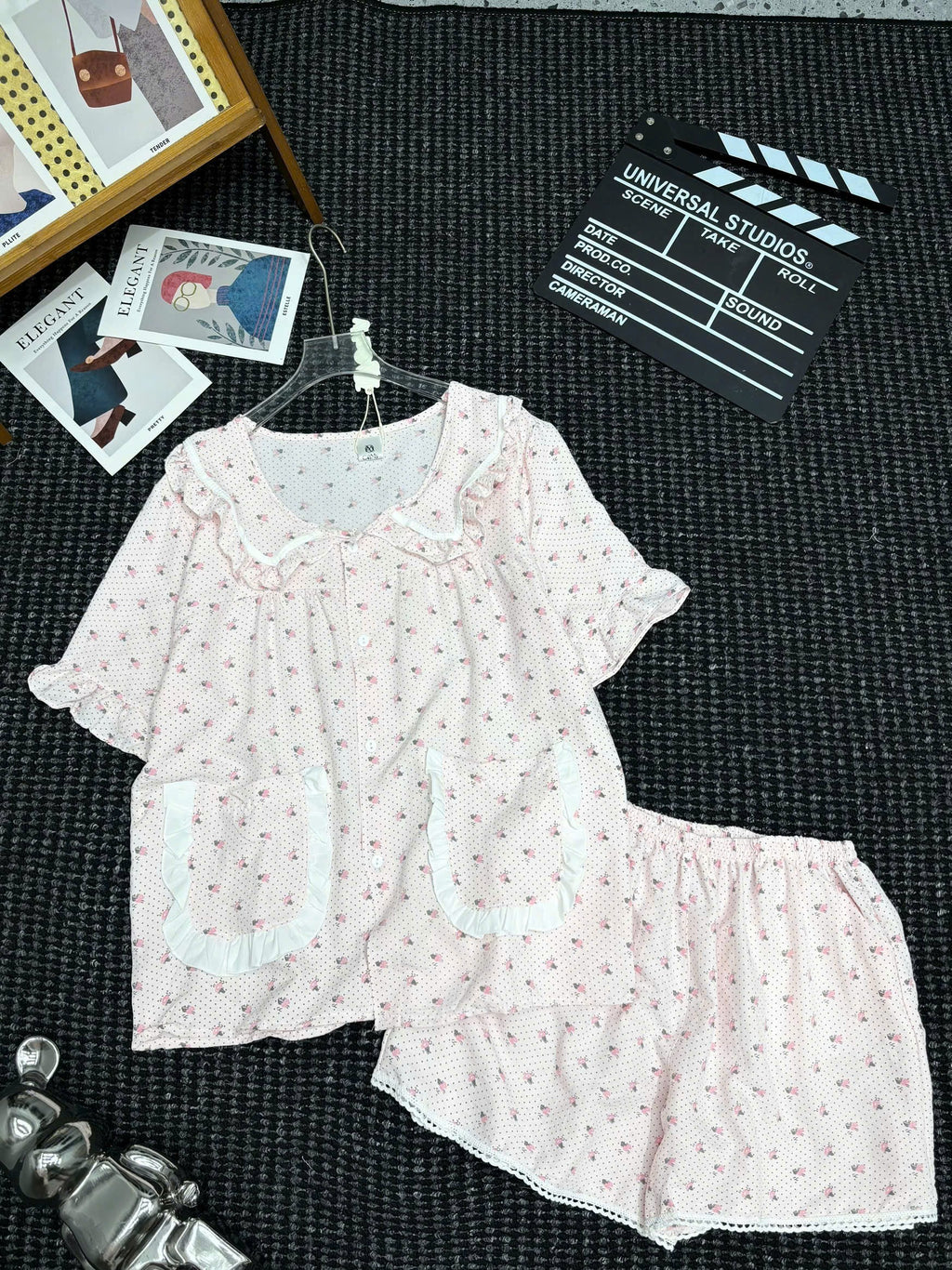 A two-piece short-sleeve pajama set laid flat on a dark textured surface, featuring a light pink top and matching shorts with tiny red floral prints and white ruffle trim. The set is styled with props including a clapperboard, cards, and a small figurine.