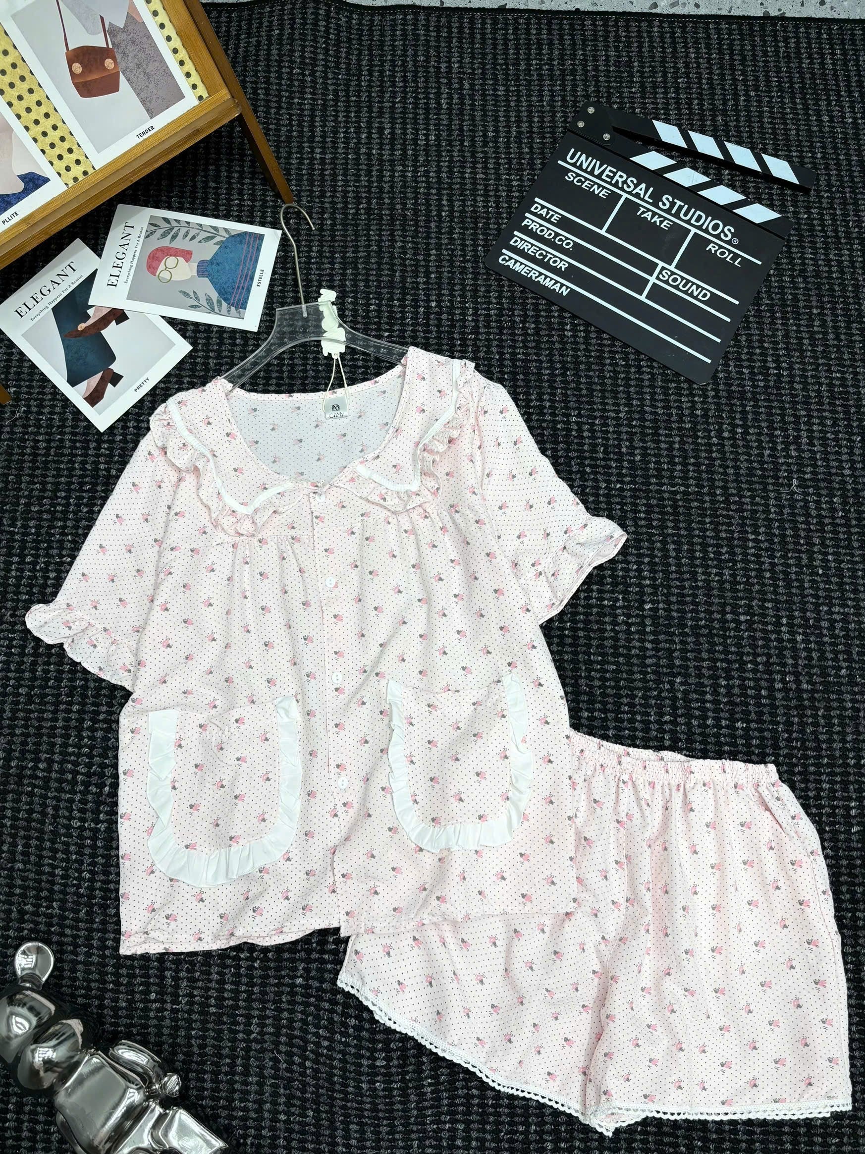 A two-piece short-sleeve pajama set laid flat on a dark textured surface, featuring a light pink top and matching shorts with tiny red floral prints and white ruffle trim. The set is styled with props including a clapperboard, cards, and a small figurine.