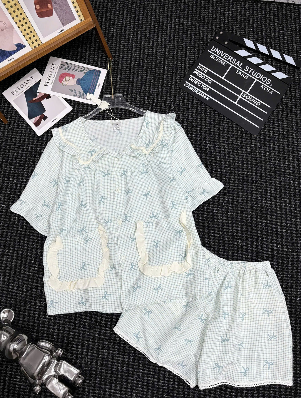 A two-piece girls' pajama set laid flat on a dark textured surface, featuring a short-sleeve top and matching shorts in a very light blue-and-white gingham pattern with small dog prints, cream ruffle trim, and a ruffled collar. Decorative props including a clapperboard, photos, and a small figurine are visible around the set.