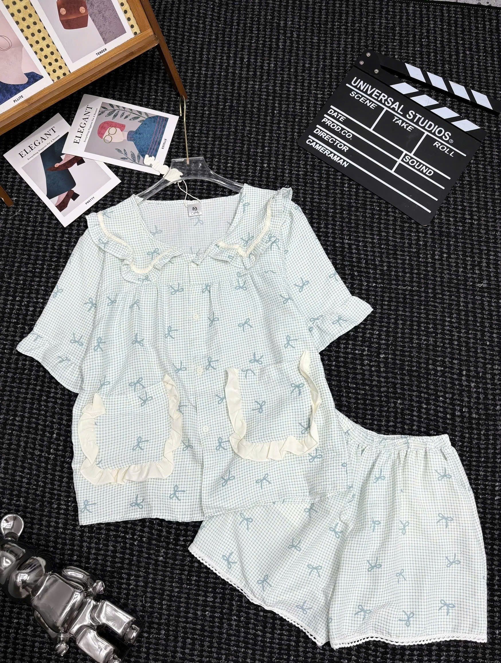 A two-piece girls' pajama set laid flat on a dark textured surface, featuring a short-sleeve top and matching shorts in a very light blue-and-white gingham pattern with small dog prints, cream ruffle trim, and a ruffled collar. Decorative props including a clapperboard, photos, and a small figurine are visible around the set.