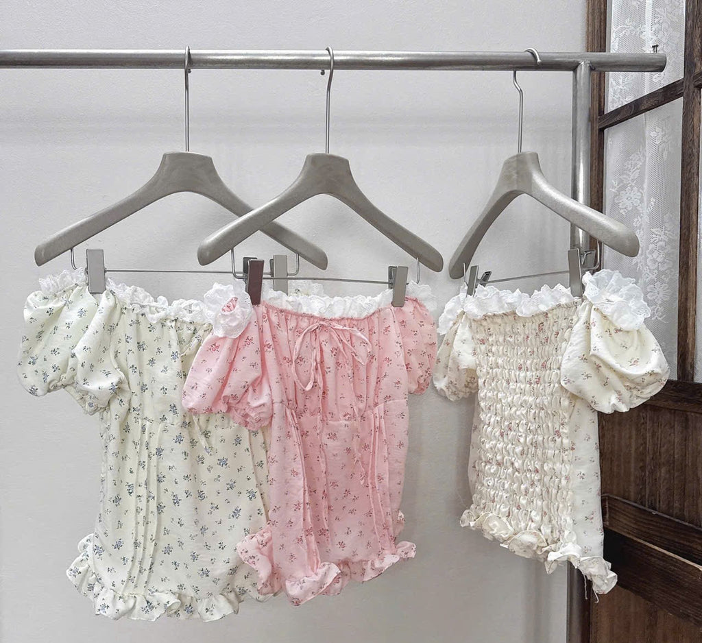 Three girls' floral and smocked romper outfits hanging on wooden hangers on a clothing rack. The outfits are cream with blue floral print, light pink with tiny blue floral print, and cream with white smocking and ruffles.