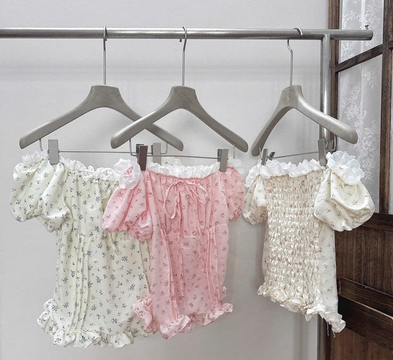 Three girls' floral and smocked romper outfits hanging on wooden hangers on a clothing rack. The outfits are cream with blue floral print, light pink with tiny blue floral print, and cream with white smocking and ruffles.
