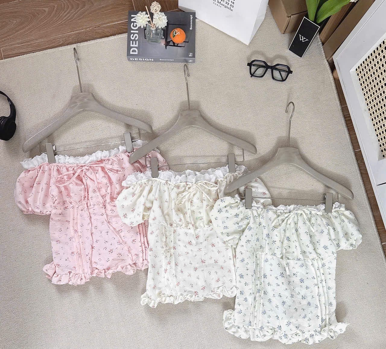 Three girl's short-sleeve romper tops with ruffled trim and tiny floral prints, displayed on wooden hangers on a beige fabric surface. The colorways are light pink, cream, and white with blue floral details.