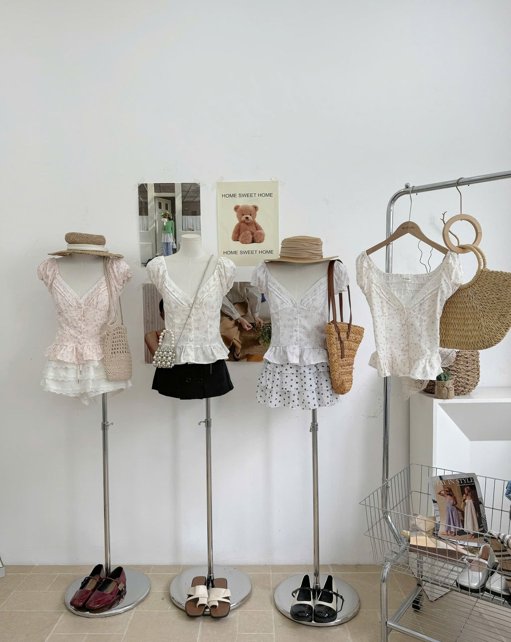 Four mannequins display women's short-sleeve tops in soft pastel colors, styled with straw hats, woven tote bags, and flats, next to a clothing rack in a bright boutique setting.