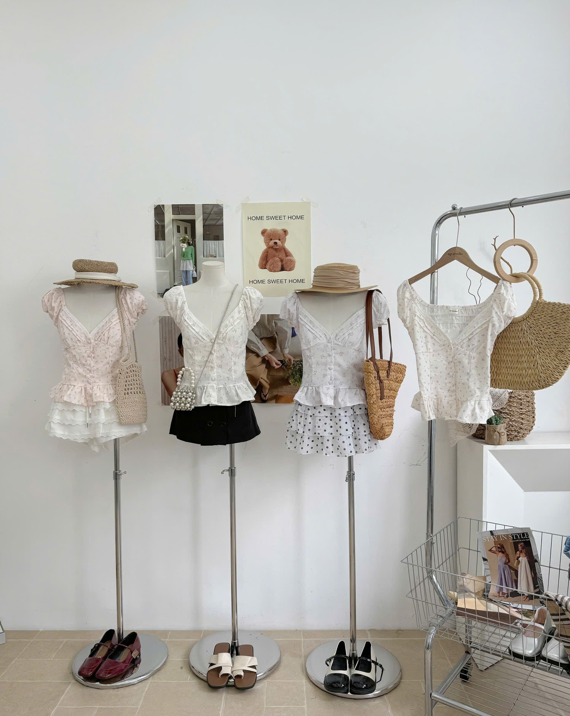 Four mannequins display women's short-sleeve tops in soft pastel colors, styled with straw hats, woven tote bags, and flats, next to a clothing rack in a bright boutique setting.