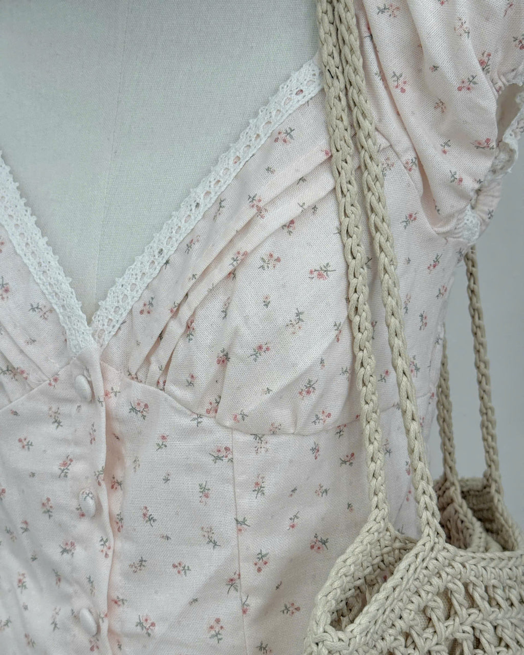 A close-up of a light blush pink floral dress with a lace V-neckline, styled on a mannequin with a beige crocheted shoulder bag.