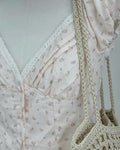 A close-up of a light blush pink floral dress with a lace V-neckline, styled on a mannequin with a beige crocheted shoulder bag.