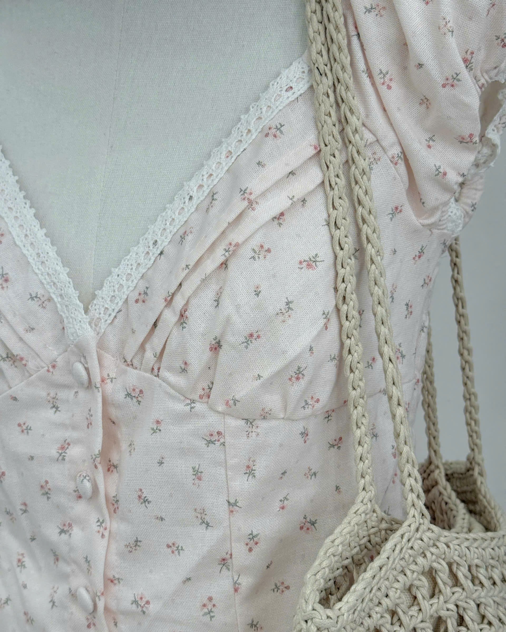 A close-up of a light blush pink floral dress with a lace V-neckline, styled on a mannequin with a beige crocheted shoulder bag.