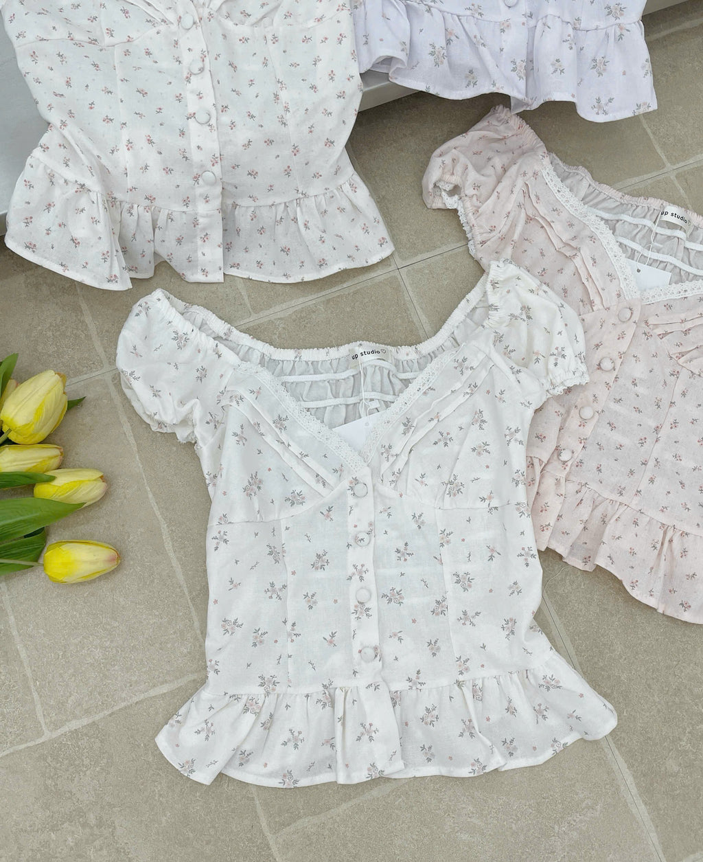 Three short-sleeve ruffled blouse tops laid out on a light stone tile floor, featuring a white floral print in the center, a white lavender print at the top left, and a pale blush pink floral print at the top right. Yellow tulips are placed on the left side.