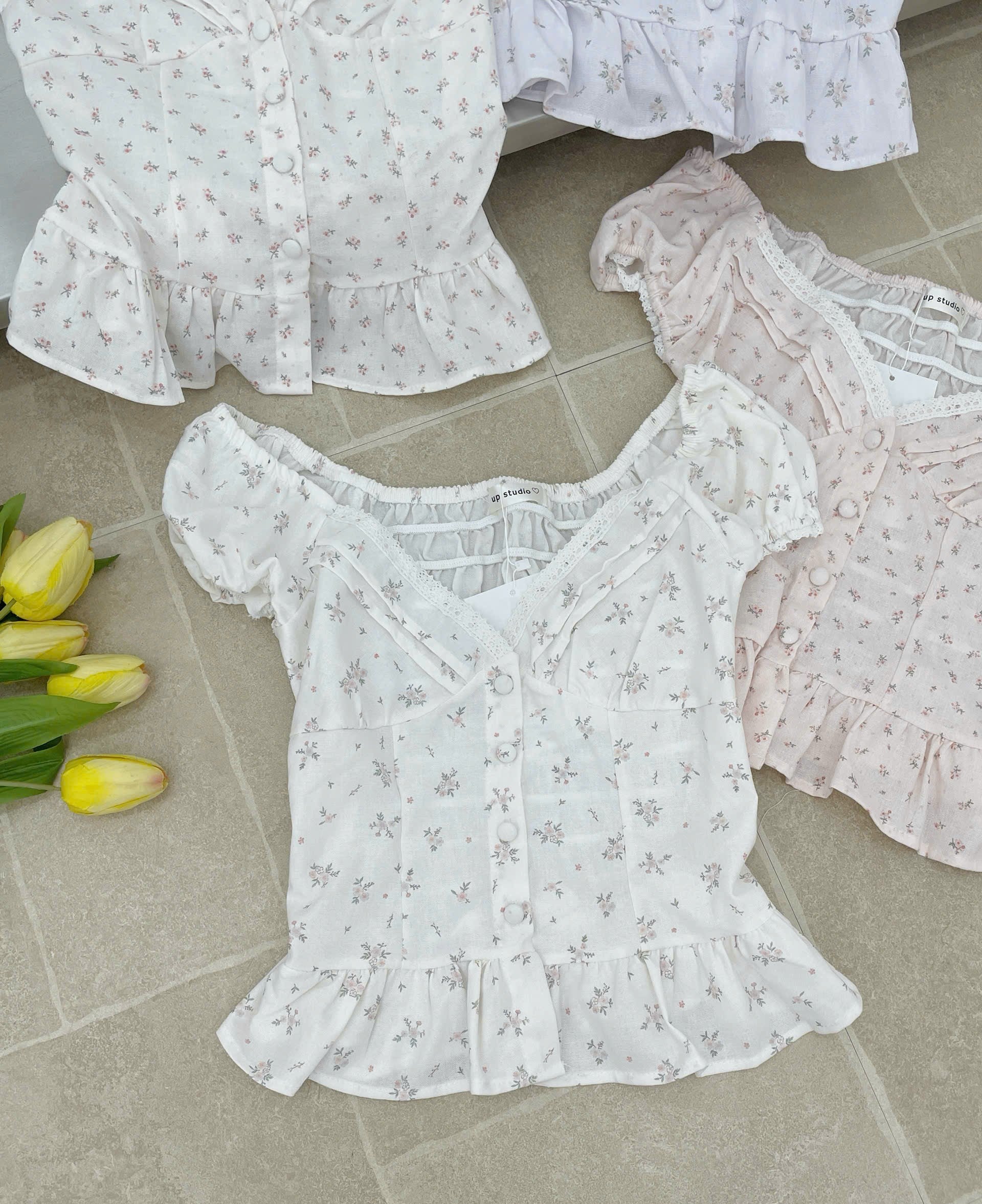 Three short-sleeve ruffled blouse tops laid out on a light stone tile floor, featuring a white floral print in the center, a white lavender print at the top left, and a pale blush pink floral print at the top right. Yellow tulips are placed on the left side.