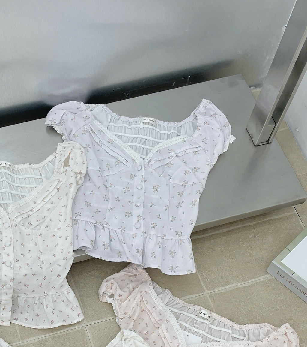 Three short-sleeve ruffled blouse tops in white, off-white, and pale pink floral prints, arranged on a gray metal shelf in a store display.