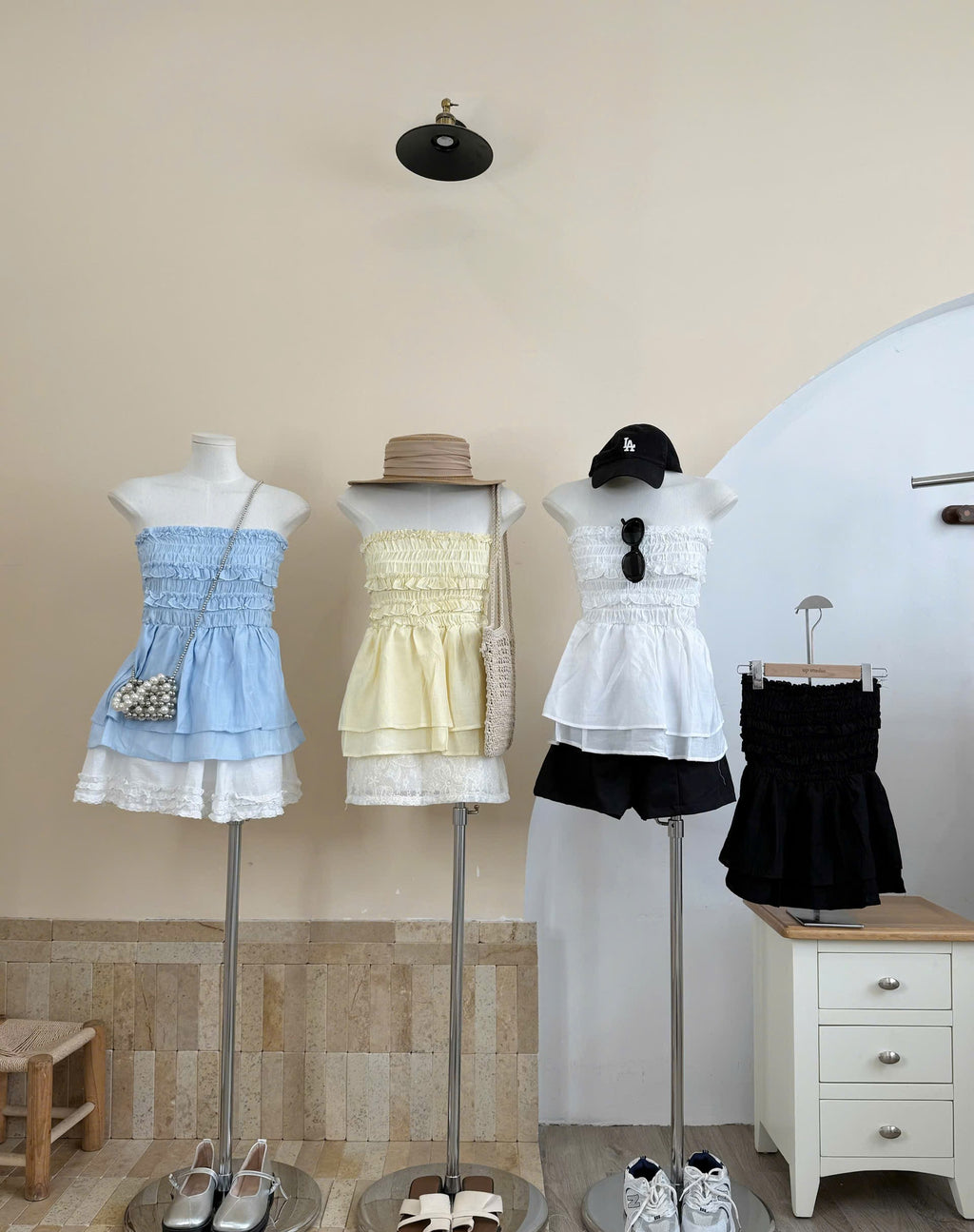 Three women's sleeveless summer outfits displayed on mannequins in a boutique setting. The left outfit is a light blue smocked ruffle dress with white lace trim and a pearl-embellished crossbody bag.