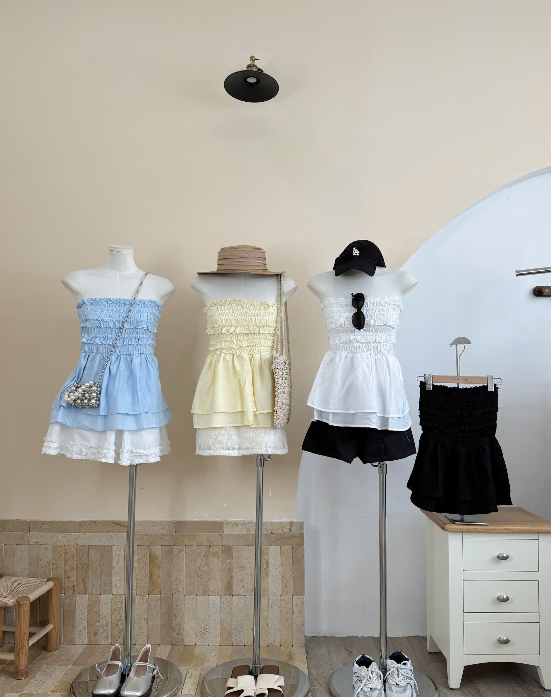 Three women's sleeveless summer outfits displayed on mannequins in a boutique setting. The left outfit is a light blue smocked ruffle dress with white lace trim and a pearl-embellished crossbody bag.
