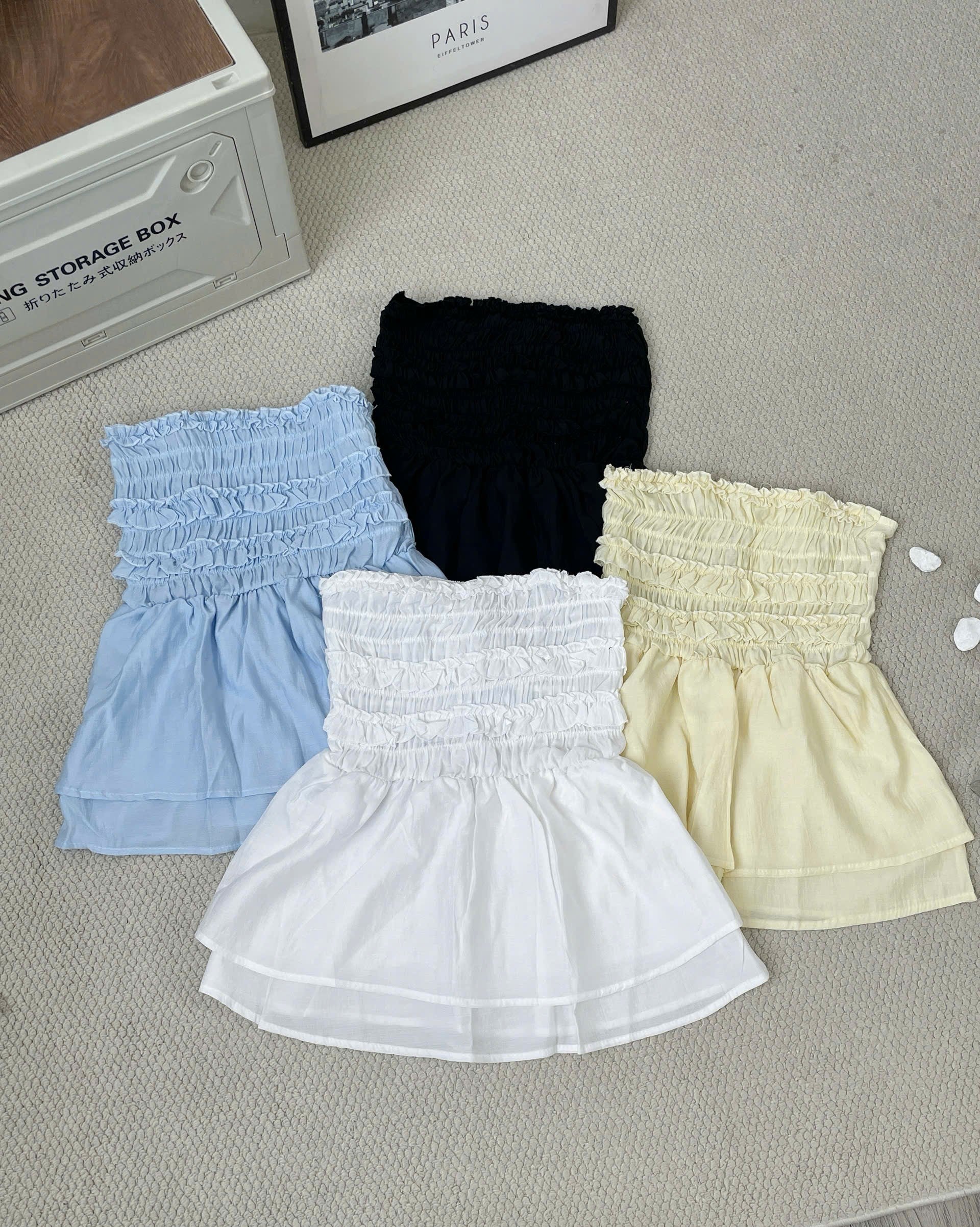 Three strapless smocked mini skirts in light blue, white, and pale yellow, arranged on a textured beige carpet with decorative props.