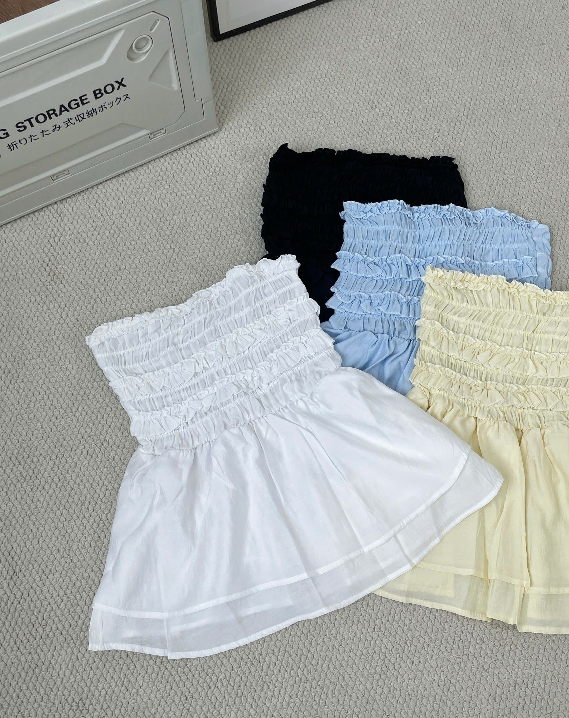 Three ruffled mini skirts in white, light blue, and pale yellow are arranged on a textured gray carpet next to a gray storage box.