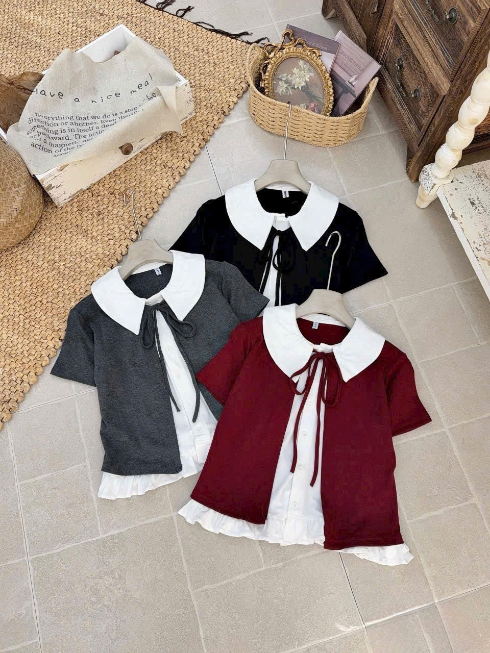 Three short-sleeve collared tops for girls displayed on hangers on a tiled floor. The set includes one dark gray top, one black top, and one burgundy top, each featuring a large white Peter Pan collar and a white ruffled undershirt peeking out at the hem.