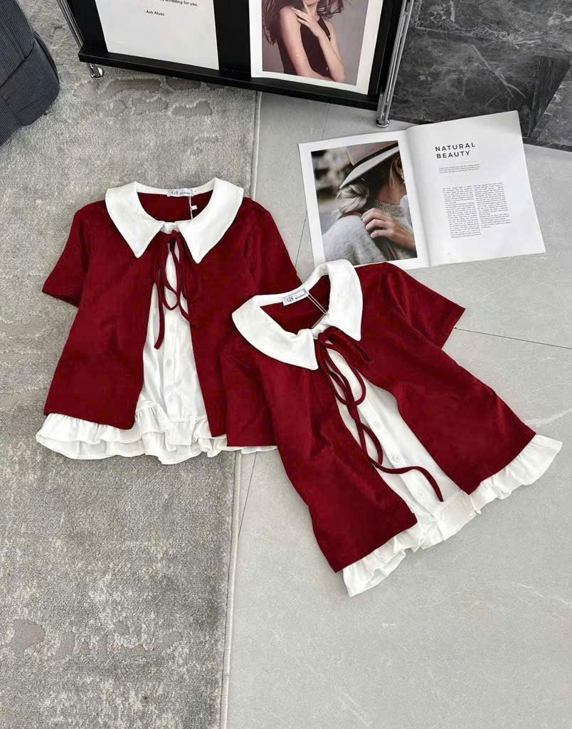 Two short-sleeve tops laid flat on a gray floor, featuring a deep red velvet-like outer layer over a white ruffled blouse with a large white collar and front tie. A magazine and framed photo are placed behind them as styling props.