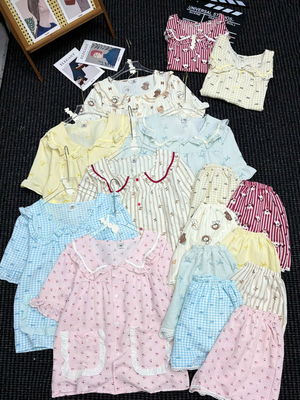 A flat lay display of multiple short-sleeve pajama sets in various colors and patterns, including pink, yellow, blue, and striped designs, arranged on a dark textured background.