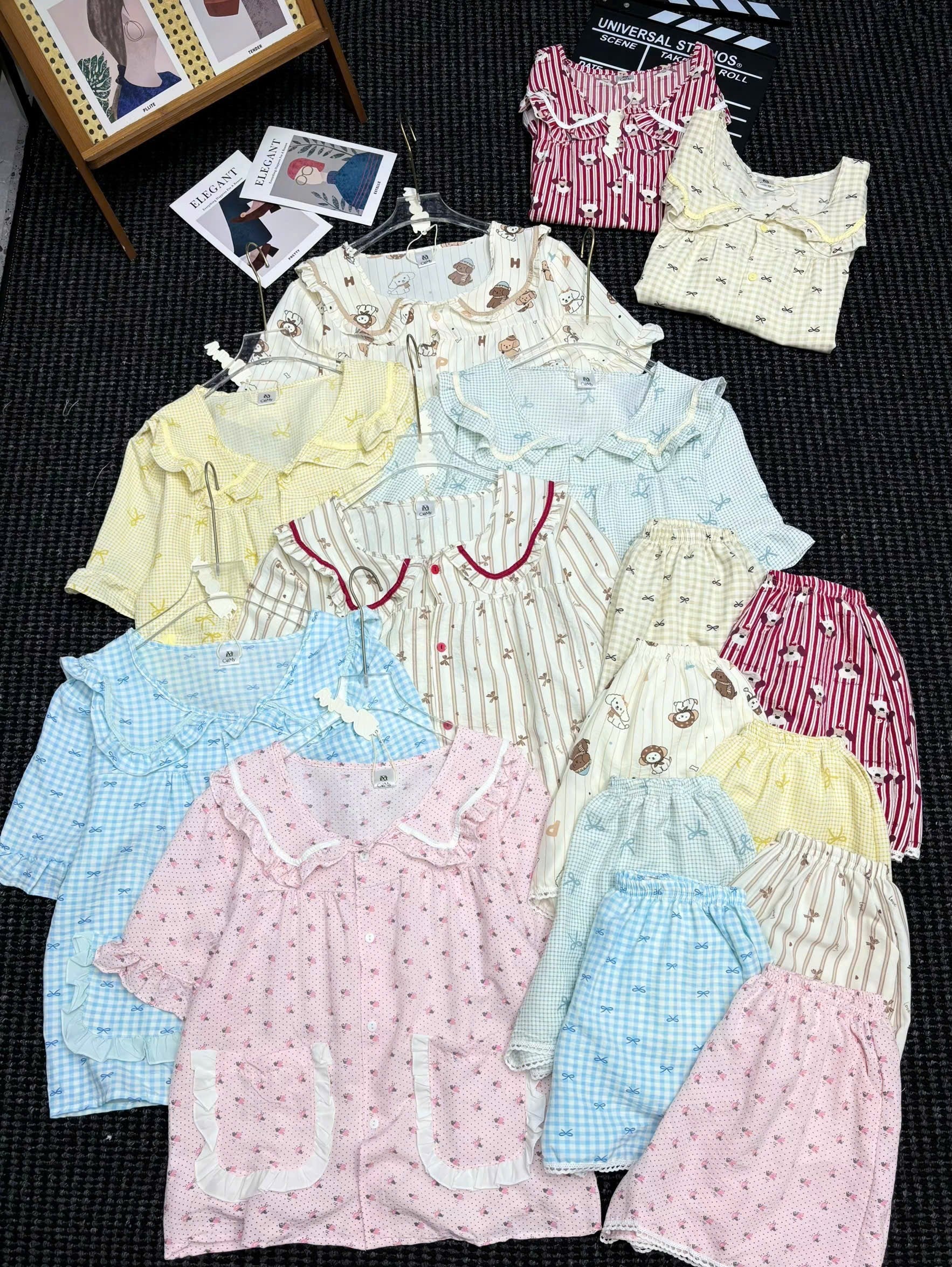A flat lay display of multiple short-sleeve pajama sets in various colors and patterns, including pink, yellow, blue, and striped designs, arranged on a dark textured background.