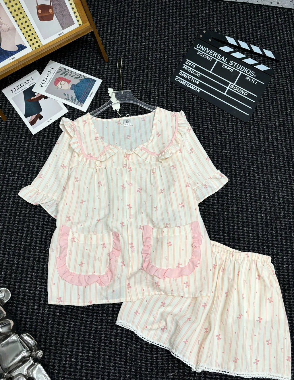 A two-piece women's pajama set laid flat on a dark textured surface, featuring a short-sleeve top and matching shorts. The pajamas have a cream and white vertical stripe pattern with small pink floral motifs, ruffled trim around the collar and pocket edges, and two front patch pockets.