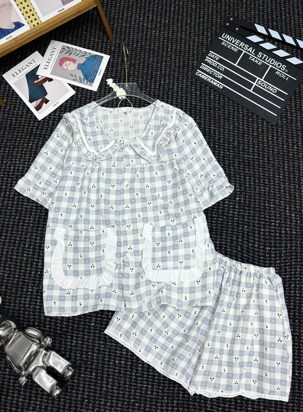 A two-piece pajama set laid flat on a dark textured surface, featuring a short-sleeve top and matching shorts in a light blue and white gingham pattern with small embroidered details and white lace trim. The set is displayed on a hanger with decorative props including a clapperboard, postcards, and a small figurine.