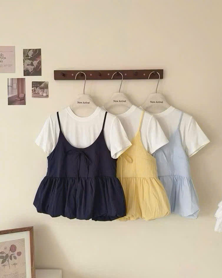 Three short-sleeve romper dresses in navy, yellow, and light blue hang on a wooden rack against a beige wall. Each romper has a white short-sleeve T-shirt layered underneath.