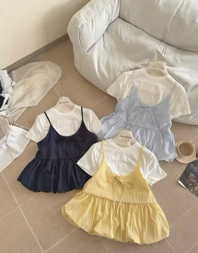 Three girl's short-sleeve dress sets styled in a flat-lay arrangement on a tiled floor next to a white sofa. The sets include a navy blue pinafore dress over a white t-shirt, a light blue pinafore dress over a white t-shirt, and a pale yellow pinafore dress over a white t-shirt.