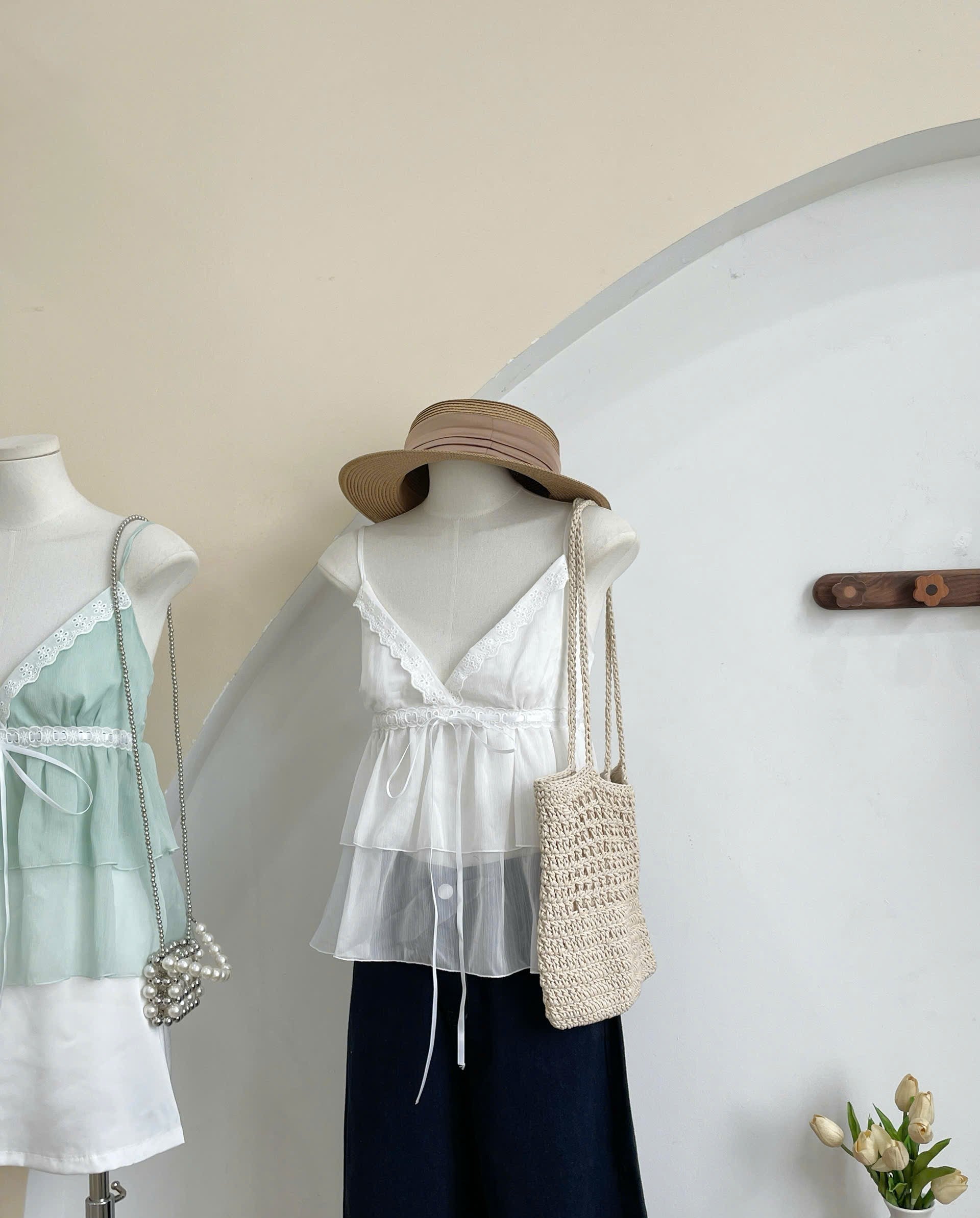 A white sleeveless tiered blouse with a sheer overlay and drawstring hem displayed on a mannequin, styled with a wide-brim tan hat, a cream woven shoulder bag, and dark navy wide-leg pants. A second mannequin in a mint green top is partially visible on the left against a cream wall with a white arched architectural feature.