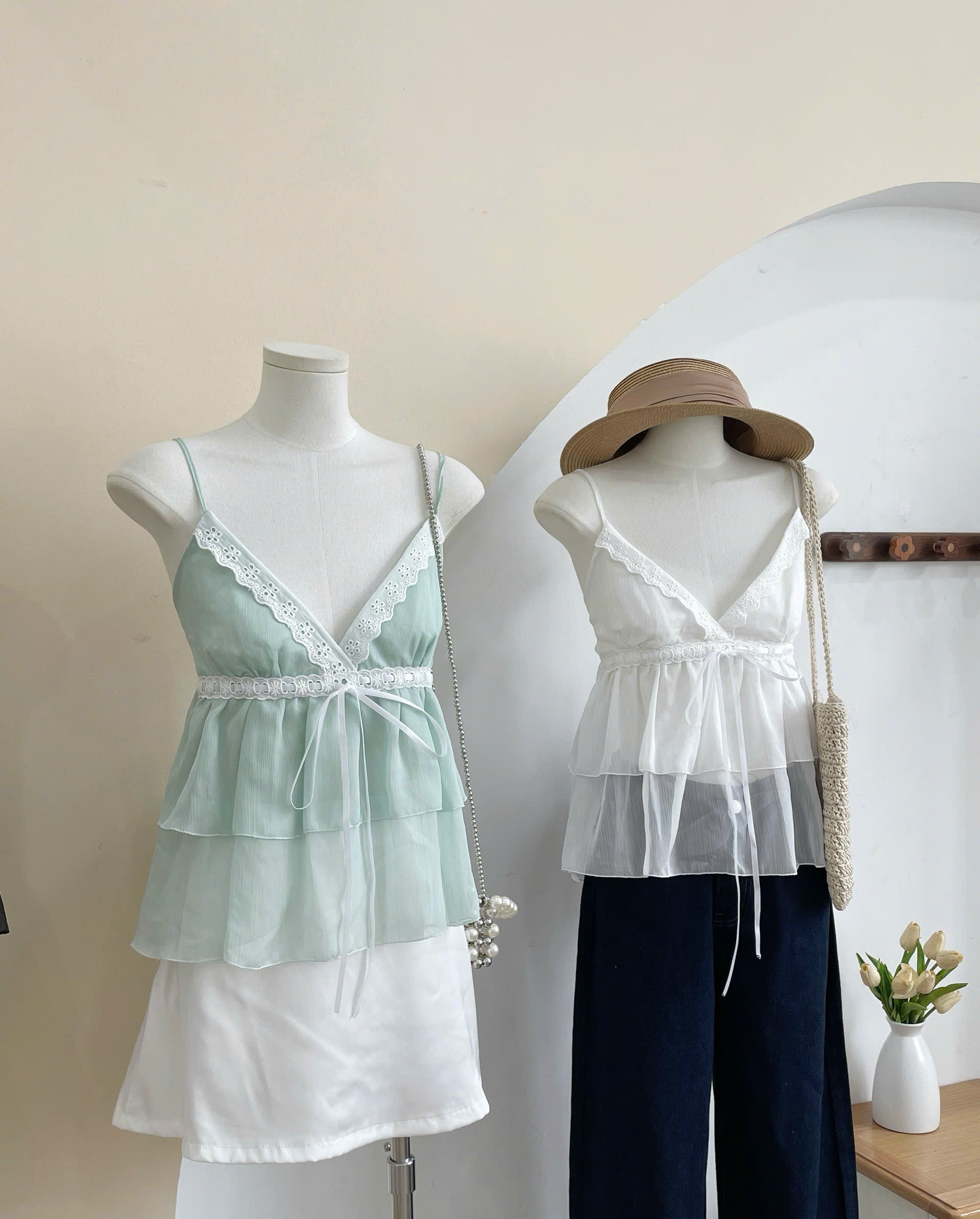 Two women's camisole-style tops displayed on mannequins indoors. The left top is a pale mint green with white lace trim and a tied waist.