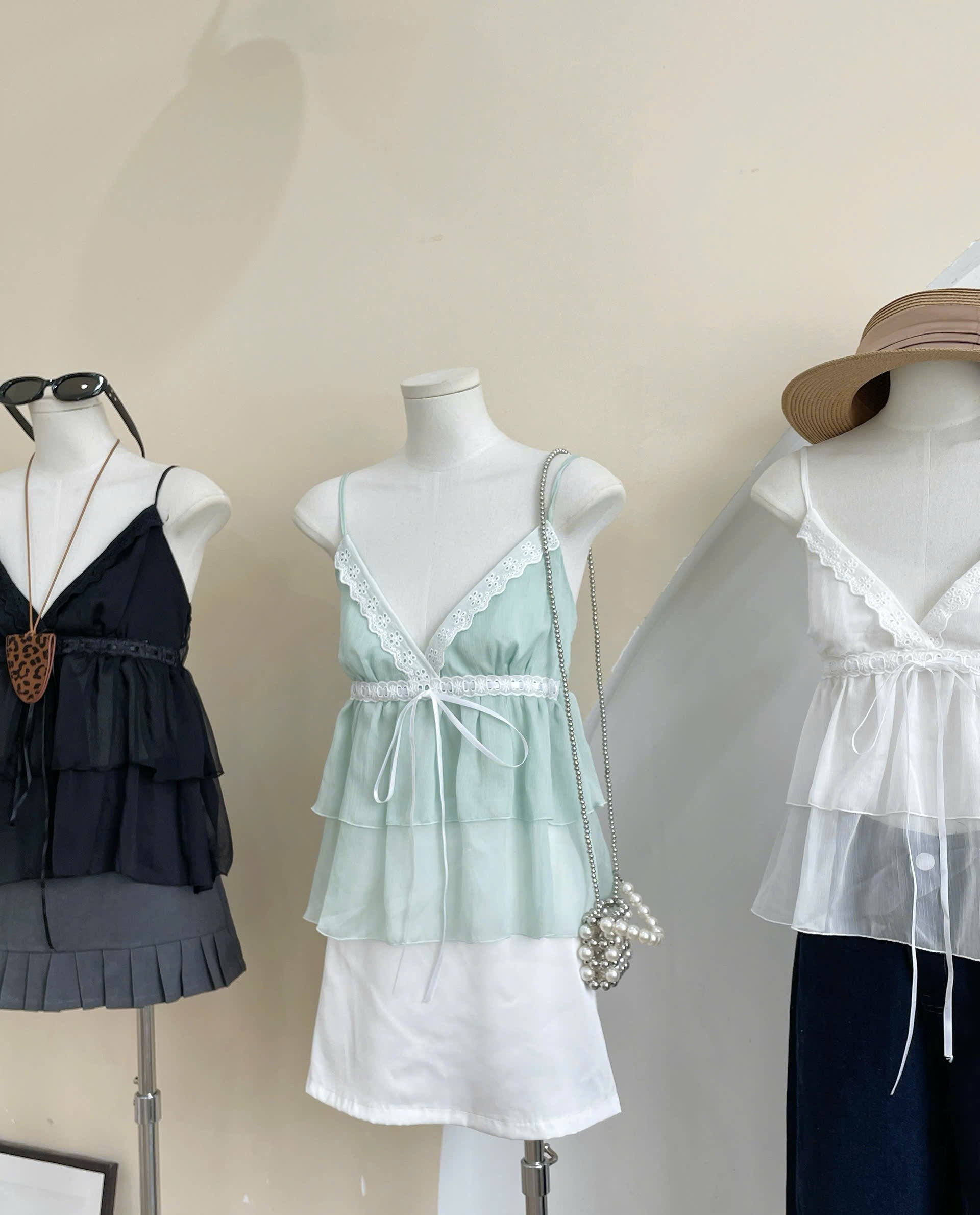Three women's camisole-style tops displayed on mannequins indoors. The center top is a pale mint green sheer ruffled camisole with white lace trim and a white underlayer.