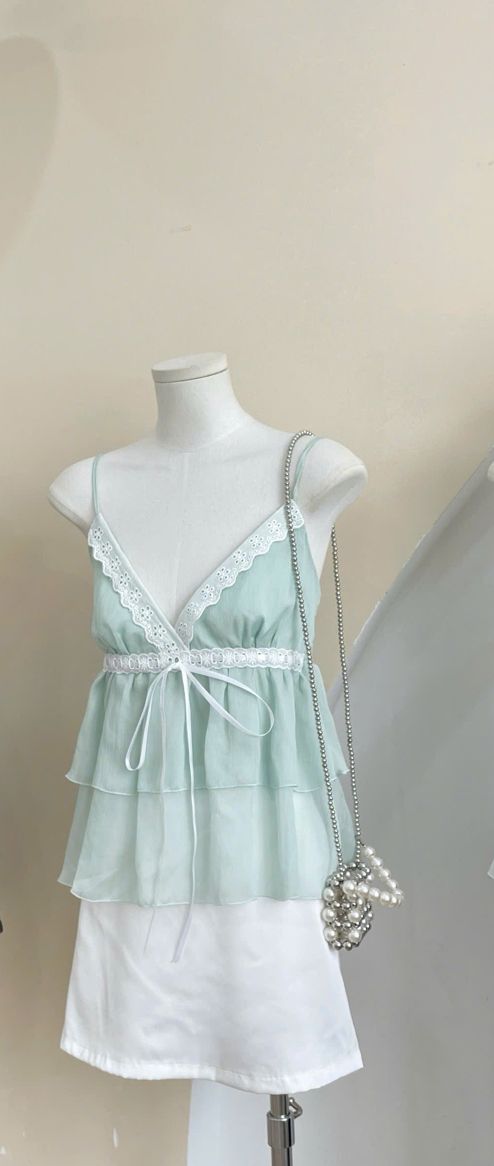A pale mint green camisole top with white lace trim and a white underskirt displayed on a mannequin, accessorized with a pearl-embellished shoulder bag.