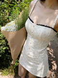 A woman outdoors wearing a cream-colored mini dress with a subtle floral pattern, black lace trim along the neckline, and ruffled straps. She is holding a large bouquet of white baby's breath in a brown paper sleeve, set against a background of green foliage and a stone path.