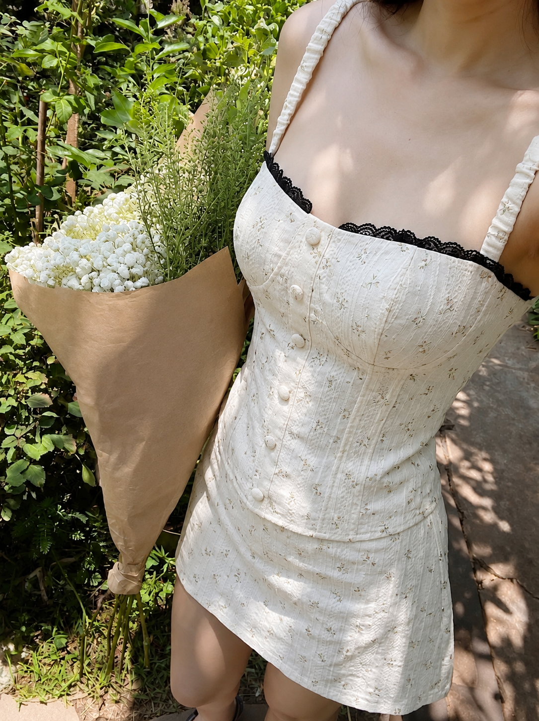 A woman outdoors wearing a cream-colored mini dress with a subtle floral pattern, black lace trim along the neckline, and ruffled straps. She is holding a large bouquet of white baby's breath in a brown paper sleeve, set against a background of green foliage and a stone path.