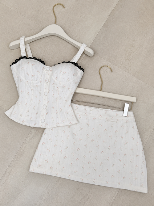 A two-piece white set consisting of a corset-style camisole top with black lace trim and a matching mini skirt, both hanging on white hangers against a beige tiled background.