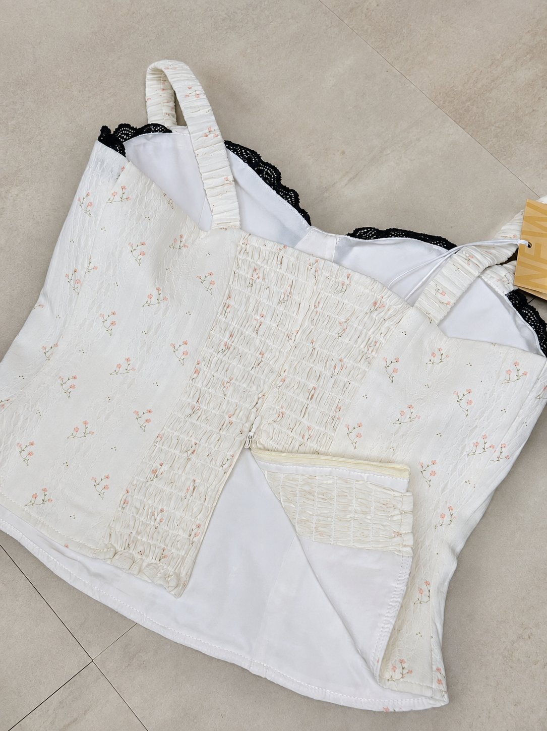 A white smocked corset-style camisole with a small floral print, black lace trim, and a white underwire bra visible underneath, laid flat on a beige tiled floor.