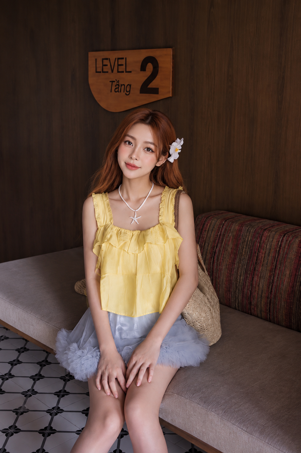 A young woman with strawberry-blonde hair sits on a gray upholstered bench against a dark wood-paneled wall. She is wearing a yellow ruffled sleeveless top and a light blue tulle skirt, with a woven straw bag resting behind her.