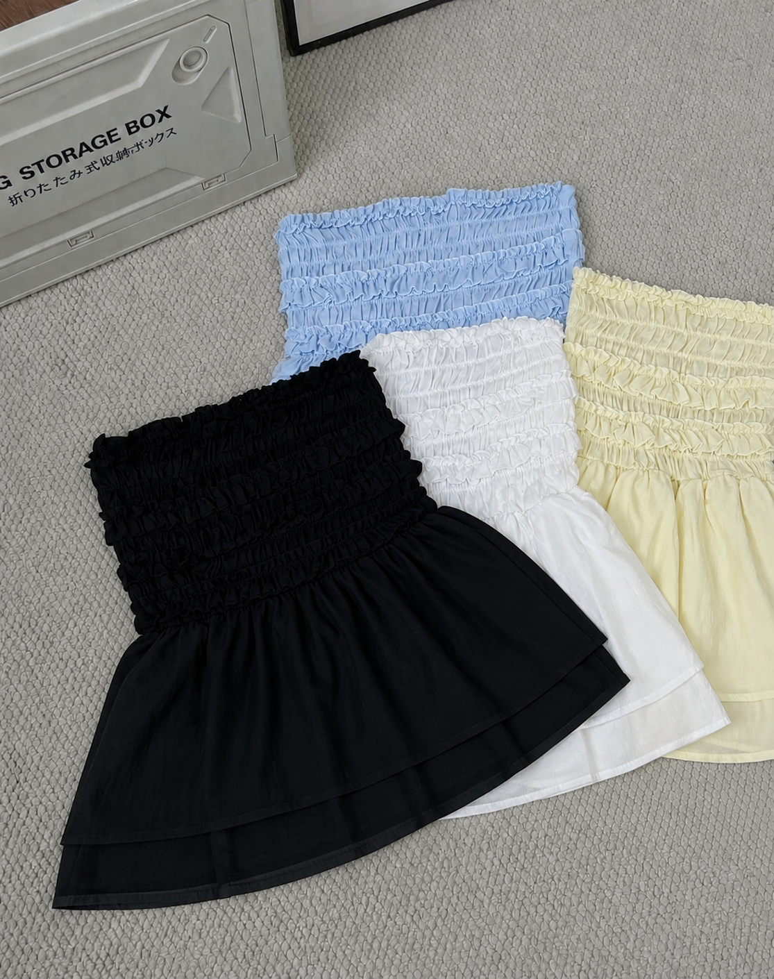 Three smocked mini skirts in black, white, and pale yellow, arranged diagonally on a gray textured carpet next to a gray storage box.