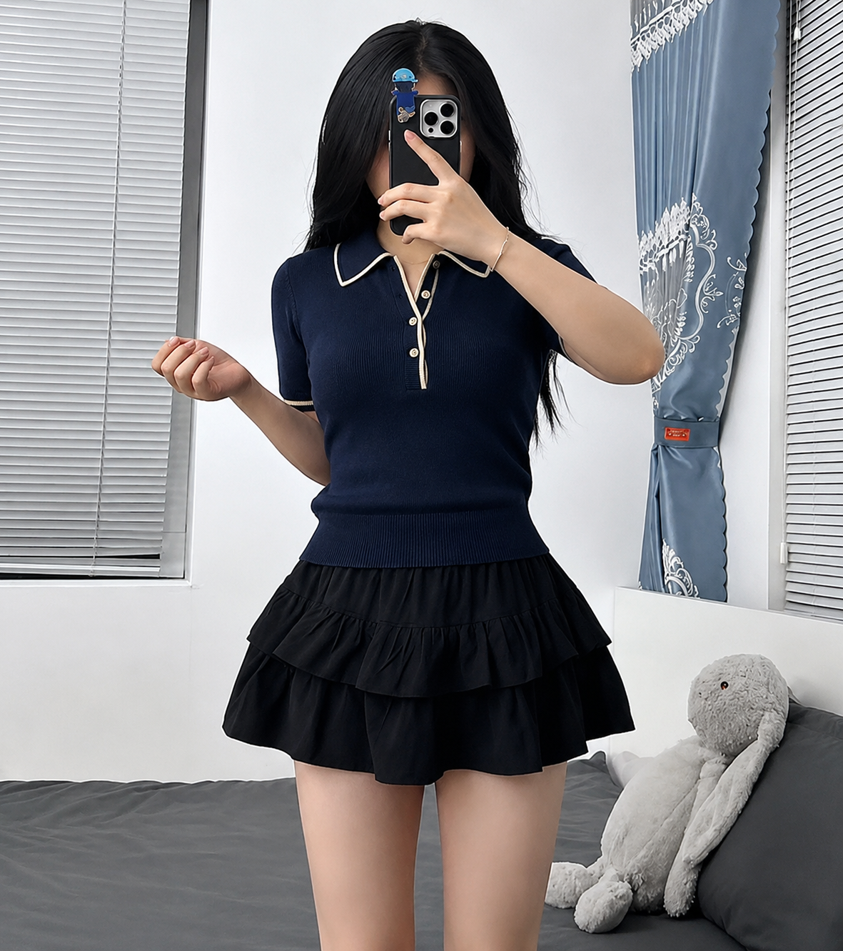 A woman takes a mirror selfie wearing a navy blue short-sleeve knit polo shirt with cream trim and a matching black tiered ruffle mini skirt. The setting is a bedroom with a gray bed, a gray stuffed bunny, and blue curtains.