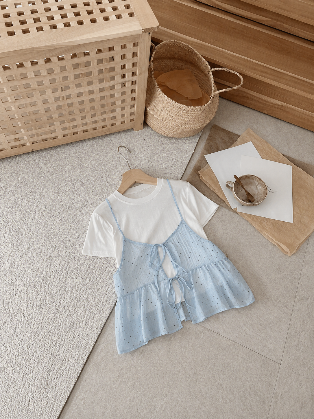A girl's two-piece outfit consisting of a white short-sleeve t-shirt and a light blue textured sleeveless romper with a tie-front detail, displayed on a wooden hanger on a light beige rug. The background features a wooden storage basket, a woven straw basket, and a wooden dresser.