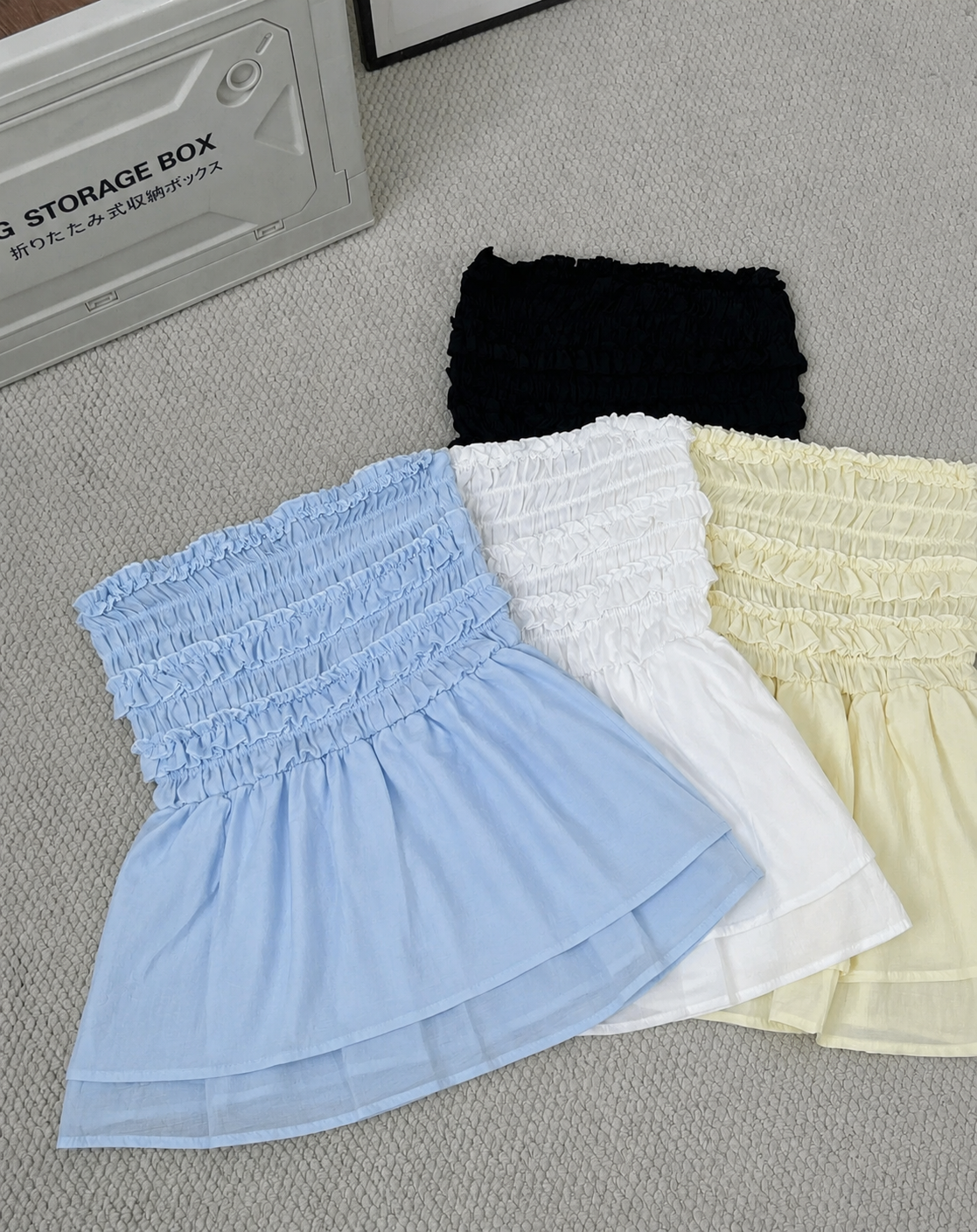 Three smocked mini skirts in light blue, white, and pale yellow are arranged on a gray textured surface next to a gray storage box.