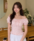 A woman wearing a light pink off-the-shoulder blouse with a small floral print, ruffled trim, and a tie at the neckline, paired with a white skirt. She is standing indoors near a wooden table and chairs.