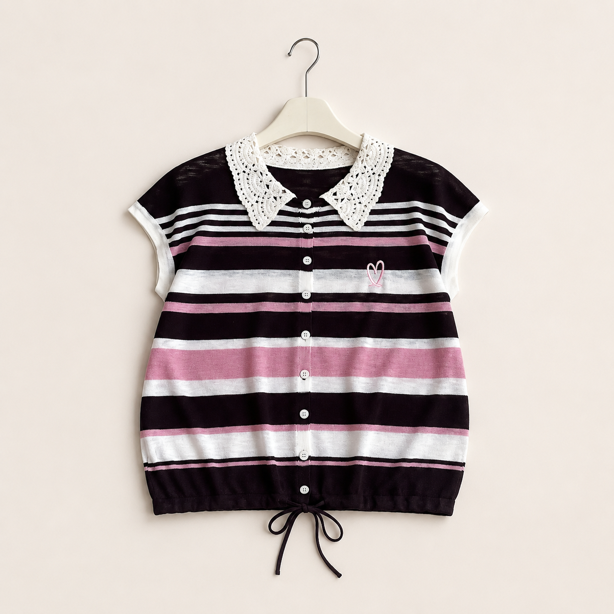 A short-sleeve knit top with horizontal stripes in black, white, and pink, featuring a white crochet collar, white buttons, and a drawstring hem, hanging on a wooden hanger against a light beige background.