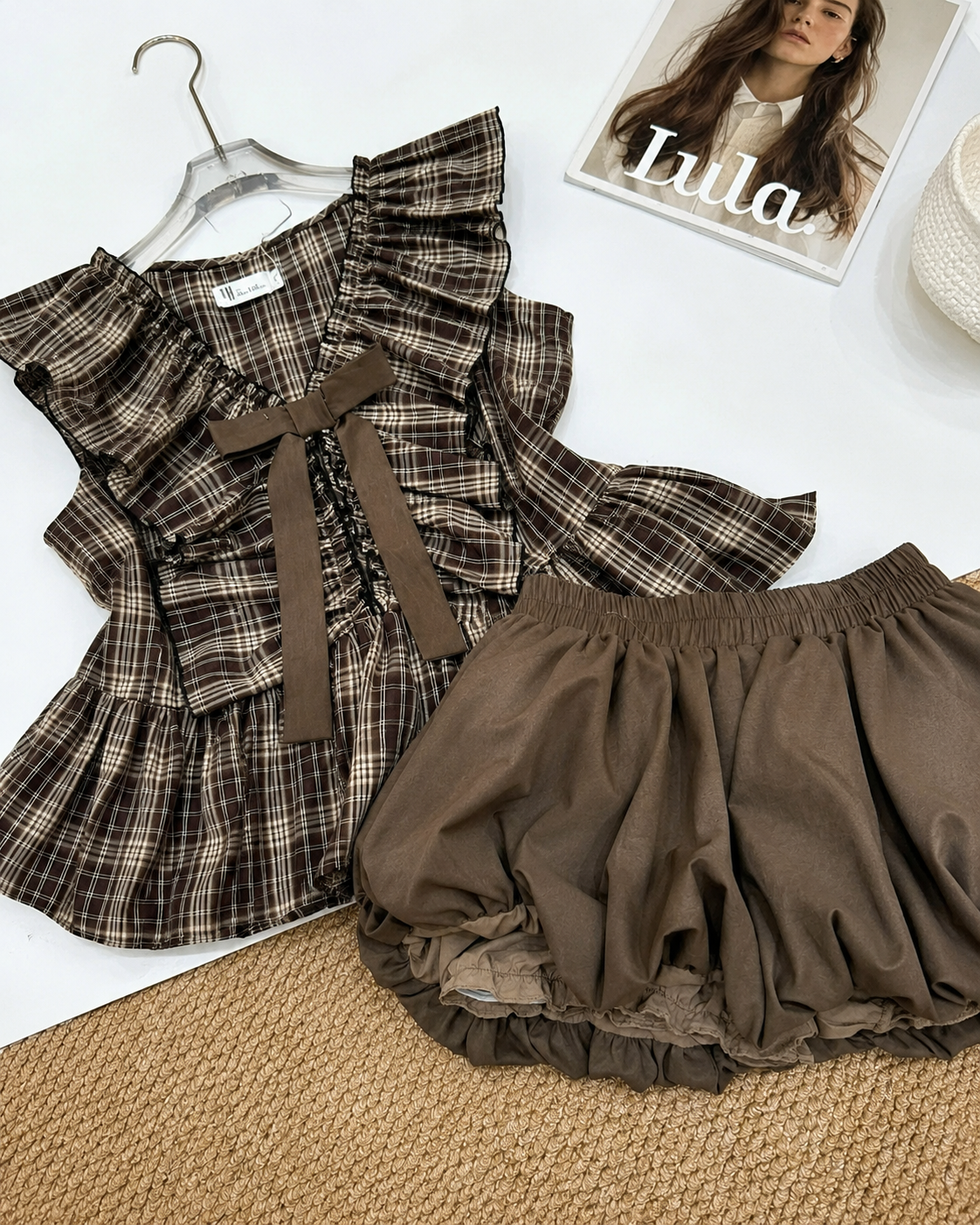 A outfit set featuring a brown and beige plaid ruffled top with a matching ribbon bow, laid flat next to matching brown ruffled shorts, styled on a white surface with a woven mat and a magazine.