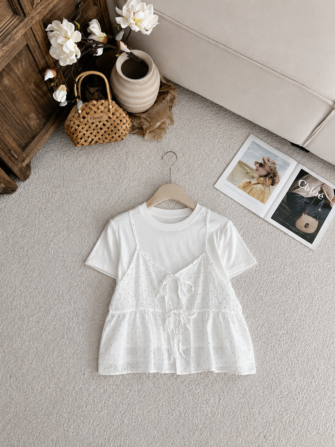 A white short-sleeve baby top with a lace applique front panel and tie detail, hanging on a wooden hanger on a light gray carpet. The background is styled with a wooden cabinet, a woven bag, a ceramic vase with flowers, and a magazine.