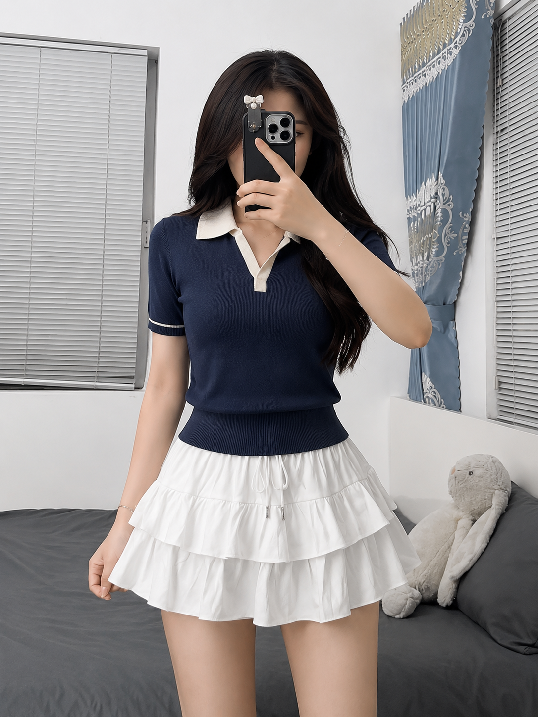 A woman takes a mirror selfie in a bedroom, wearing a navy blue short-sleeve polo knit top with a cream collar and a white tiered ruffle mini skirt.
