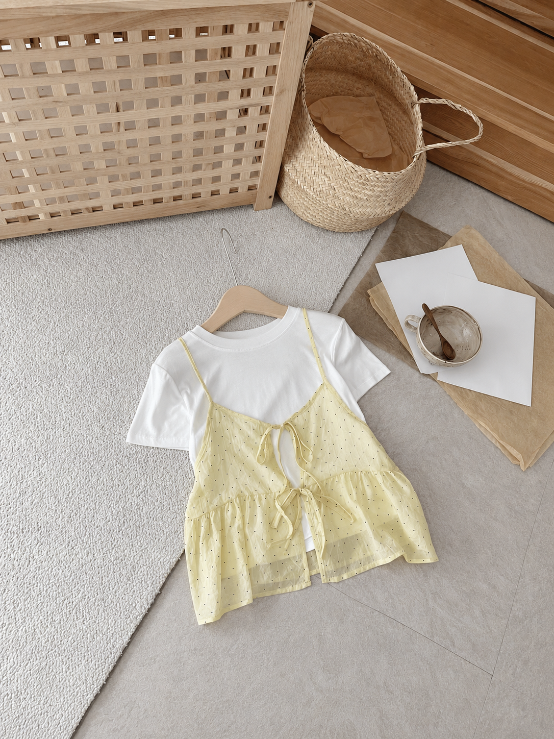 A two-piece outfit set featuring a white short-sleeve t-shirt and a pale yellow dotted camisole dress, styled on a light wood hanger on a gray rug with neutral props.